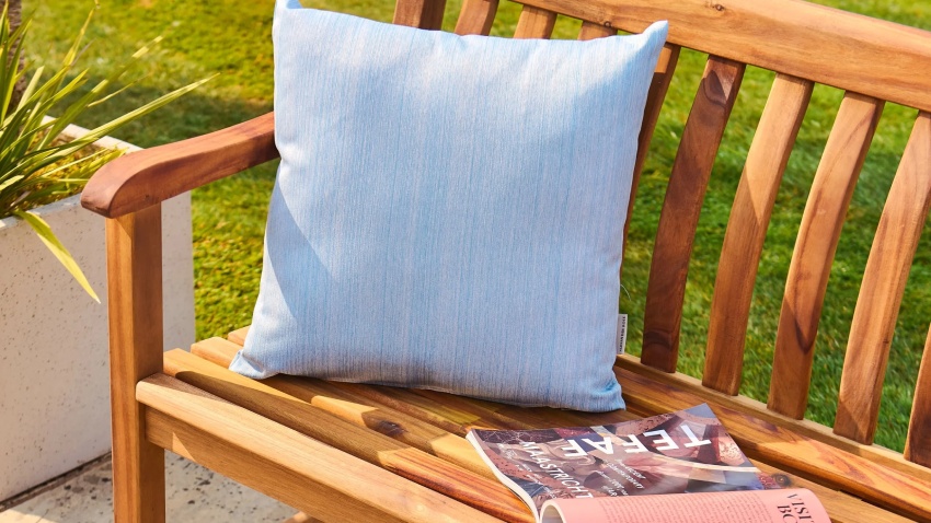 Close-up of Alexander Rose Redcliffe Broadfield Bench 5ft 335R in acacia wood, showing slatted seat, curved armrest, and blue outdoor cushion in a sunlit garden setting
