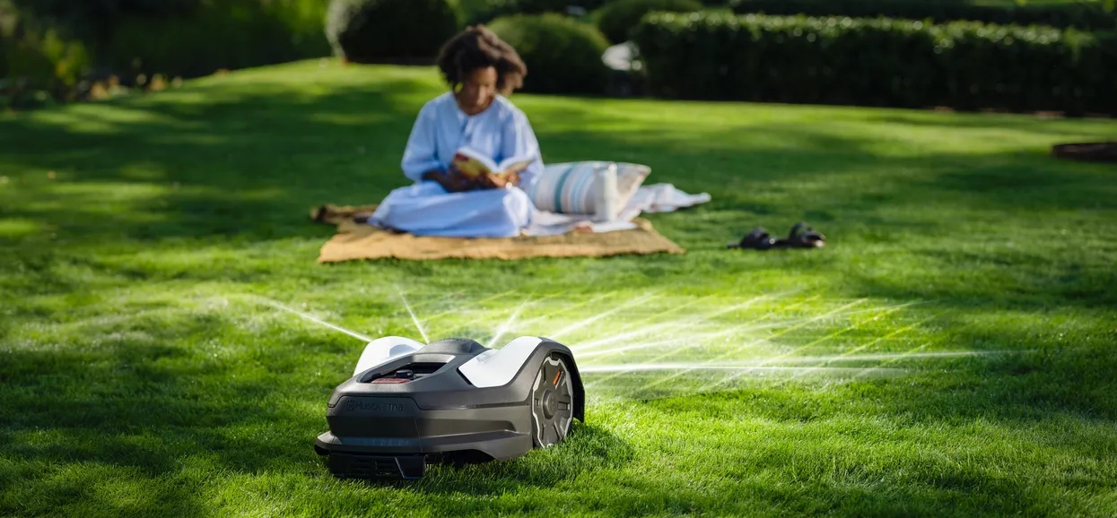 Husqvarna Automower® 405VE NERA robotic lawn mower using AI vision to navigate a garden lawn, operating wire-free while a homeowner relaxes nearby.