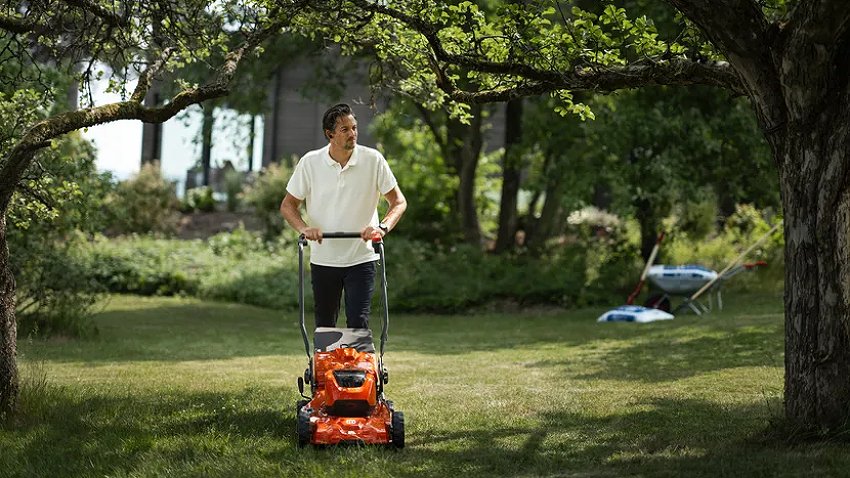 Person using a Husqvarna LC251iS self-propelled battery lawnmower to mow a garden lawn beneath mature trees. Cordless mower designed for efficient mowing of medium to large gardens.