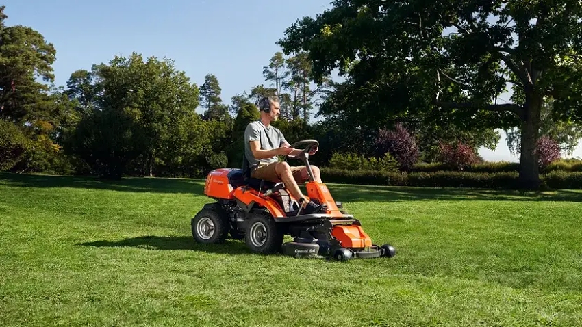 Husqvarna R214T ride-on lawnmower in use on a large lawn, featuring a front-mounted cutting deck for precise mowing around obstacles and landscaped gardens.