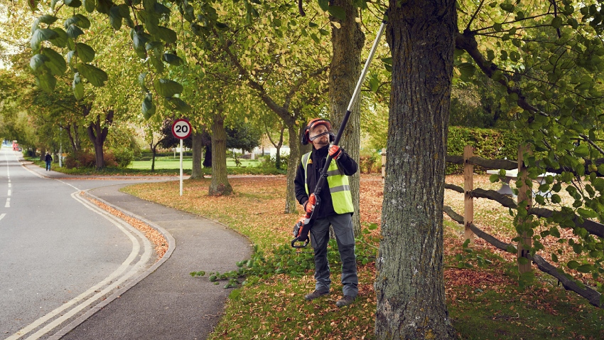 Husqvarna 530iPT5 battery telescopic pole saw in use cutting tree branches roadside, professional operator wearing protective gear