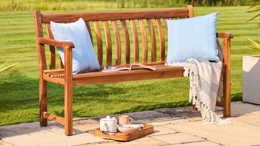 Alexander Rose Redcliffe Broadfield Bench 5ft 335R in acacia wood, styled with cushions and throw on a patio in a manicured garden, full bench view