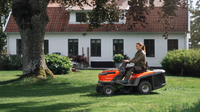 A woman using the Husqvarna TC100i ride-on garden tractor mower cutting a large lawn with battery-powered collector mower in residential garden