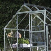 Close-up of the Halls ICON 6 6x10 toughened glass greenhouse with a natural mill-finish aluminium frame. Door slid open to show integrated staging.