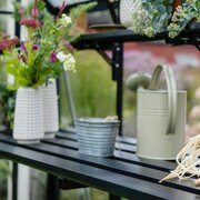 Halls modular folding greenhouse staging installed inside an ICON greenhouse, showing the black slatted shelf holding plant pots and a metal watering can.