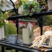 Halls modular folding staging HR01913 installed inside an ICON 8 8x14 greenhouse, showing the black slatted shelf holding plant pots, flowers, and a metal watering can.