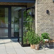 Side view of the Halls QUBE 42 4x2 black lean-to greenhouse with toughened glass panels, positioned next to bi-fold doors with patio plants and shelving inside.