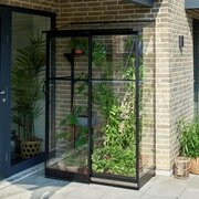 Angled view of the Halls QUBE 42 4x2 lean-to greenhouse in black aluminium with toughened glass, showing interior shelving, climbing plants and patio seating nearby.