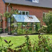 Full-length side view of the Halls QUBE 6x10 greenhouse in black toughened glass installed beside a brick house, filled with plants and growing vegetables.