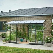 Front view of the Halls QUBE 612 6x12 lean-to greenhouse with black aluminium frame and toughened glass, filled with plants and accessories against a brick wall.