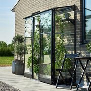Angled view of the Halls QUBE 62 6x2 lean-to greenhouse in black with toughened glass, installed against a brick wall with sliding doors, compact growing space, and potted plants on a modern patio.