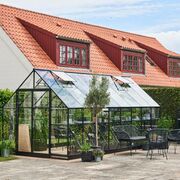 Wide patio view of the Halls Qube+ 816 8x16 black aluminium greenhouse with toughened glass roof and sides, showing outdoor dining furniture, internal staging, and the full elongated footprint.