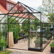 Angled front view of the Halls Qube+ 816 8x16 black toughened glass greenhouse showing low-threshold double doors, roof vent open, internal metal staging, potted plants, and walkway access on a brick base.