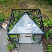 A top-down view of an 8x8 black-framed Halls Qube greenhouse filled with plants, showing its glass roof structure and paved base surrounded by garden beds.