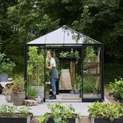 A gardener tending plants inside a black-framed Halls Qube 8x8 greenhouse with toughened glass, surrounded by raised beds and outdoor greenery.