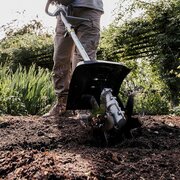 Gardener using the Honda SSCL VersaTool Cultivator Attachment to break up soil in a flower bed, showing the rotating tines working through compacted earth in a garden setting.