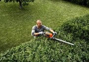 Husqvarna 215iHD45 battery hedge trimmer in use cutting a mature garden hedge, featuring a 45cm double-sided blade for precise hedge maintenance.