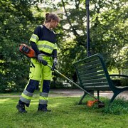 Operator using Husqvarna 525iLXT cordless battery trimmer to cut grass around garden bench, precise edging and trimming work