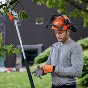 Operator preparing to use Husqvarna 530iP4 battery pole saw, wearing safety helmet and gloves, pruning trees in garden