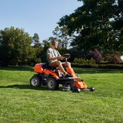 Person operating a Husqvarna R214T ride-on lawnmower on a large lawn. Front-mounted cutting deck provides precise mowing around trees, borders and landscaped garden areas.