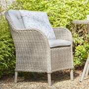 Leisure Grow St Tropez Sand garden dining chair with woven rattan-effect frame and light grey seat and back cushions, shown on a gravel patio.