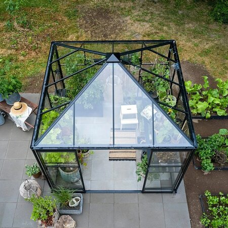 A top-down view of an 8x8 black-framed Halls Qube Square greenhouse installed professionally and filled with plants. The glass roof structure and paved base can be seen, surrounded by garden beds.
